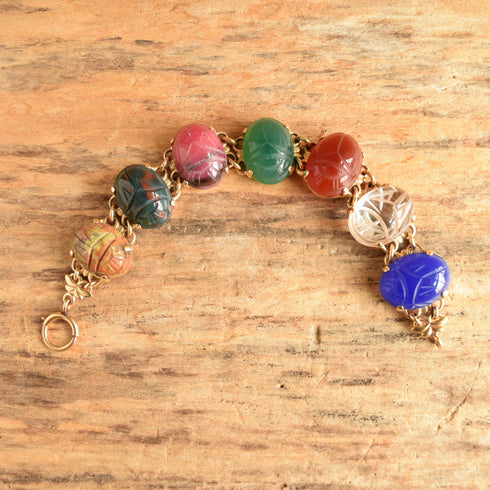 A 14k yellow gold link bracelet featuring 7 carved oval gemstone scarab beetles on a wooden background.
