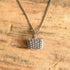 A silver-toned chain necklace with a 3d picnic basket shaped pendant, displayed on a textured tan stone surface.
