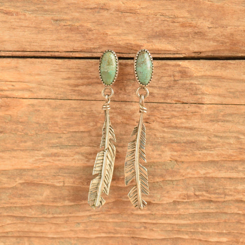A pair of silver-toned drop earrings with oval green turquoise-like stones at the top and feather-shaped dangling elements below, displayed against a rustic wooden surface.
