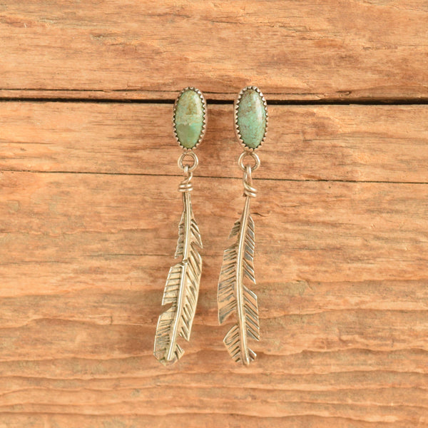 A pair of silver-toned drop earrings with oval green turquoise-like stones at the top and feather-shaped dangling elements below, displayed against a rustic wooden surface.
