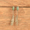 A pair of silver-toned drop earrings with oval green turquoise-like stones at the top and feather-shaped dangling elements below, displayed against a rustic wooden surface.