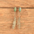 A pair of silver-toned drop earrings with oval green turquoise-like stones at the top and feather-shaped dangling elements below, displayed against a rustic wooden surface.