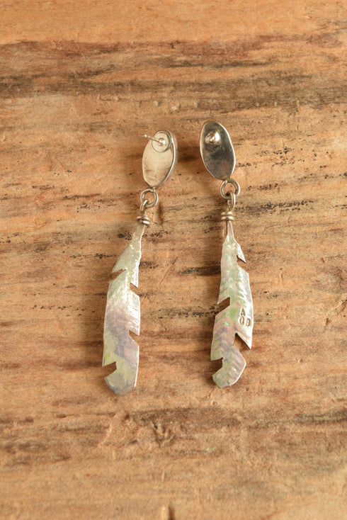 A pair of silver-toned dangle earrings with feather-shaped charms and oval drop charms, displayed on a textured brown wooden surface.