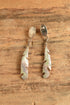 A pair of silver-toned dangle earrings with feather-shaped charms and oval drop charms, displayed on a textured brown wooden surface.