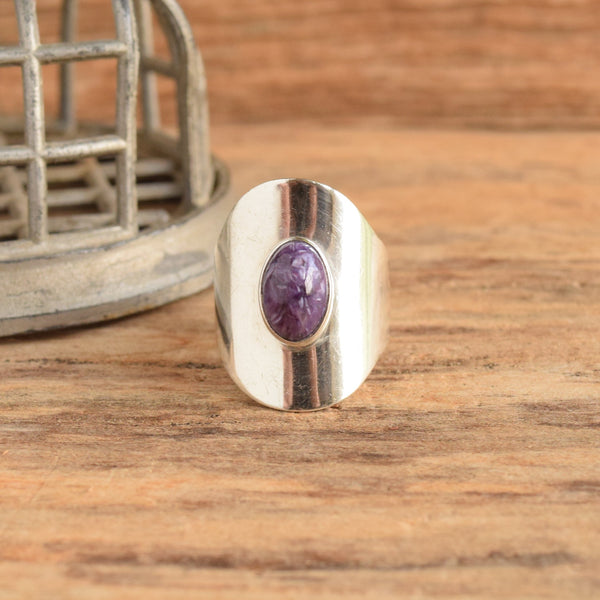 A silver-toned ring with a central oval purple gemstone, displayed on a rustic wooden surface next to a small metal basket.