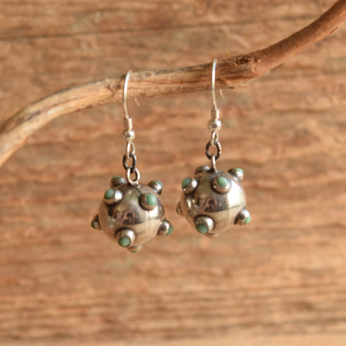 A pair of sterling silver dangle earrings with small green stone accents, hanging from a natural branch against a rustic wooden background.