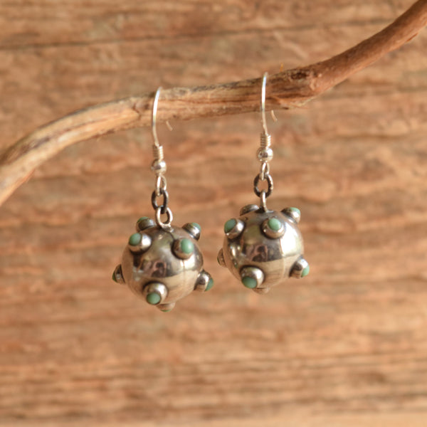 A pair of sterling silver dangle earrings with small green stone accents, hanging from a natural branch against a rustic wooden background.