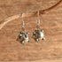 A pair of sterling silver dangle earrings with small green stone accents, hanging from a natural branch against a rustic wooden background.