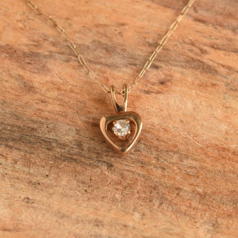 A delicate gold necklace with a heart-shaped pendant featuring a small clear gemstone, displayed on a textured brown wooden surface.