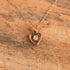 A delicate gold necklace with a heart-shaped pendant featuring a small clear gemstone, displayed on a textured brown wooden surface.