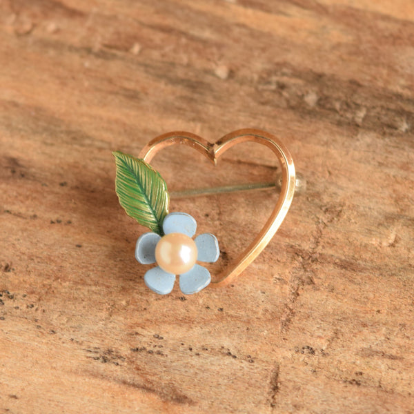 A small gold heart-shaped ring with a light blue flower and green leaf pin, resting on a textured wooden surface.