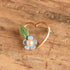 A small gold heart-shaped ring with a light blue flower and green leaf pin, resting on a textured wooden surface.