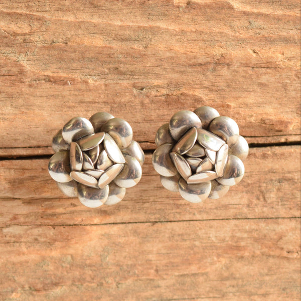 A pair of silver-toned flower shaped stud earrings resting on a rustic, textured wooden surface.