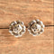 A pair of silver-toned flower shaped stud earrings resting on a rustic, textured wooden surface.