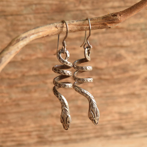 A pair of silver snake dangle earrings hanging from a natural branch against a rustic wooden background.