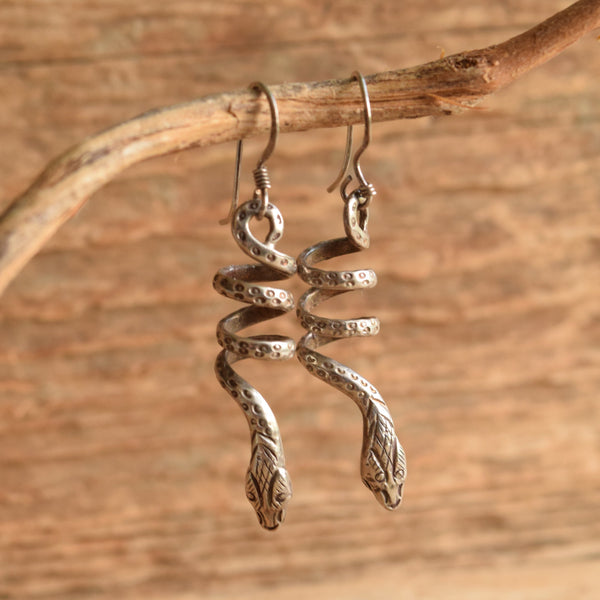 A pair of silver snake dangle earrings hanging from a natural branch against a rustic wooden background.