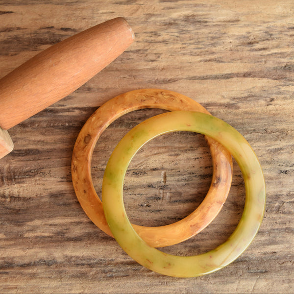 Two Bakelite flying saucer bangle bracelets in brown and green colors, displayed on a wooden surface.